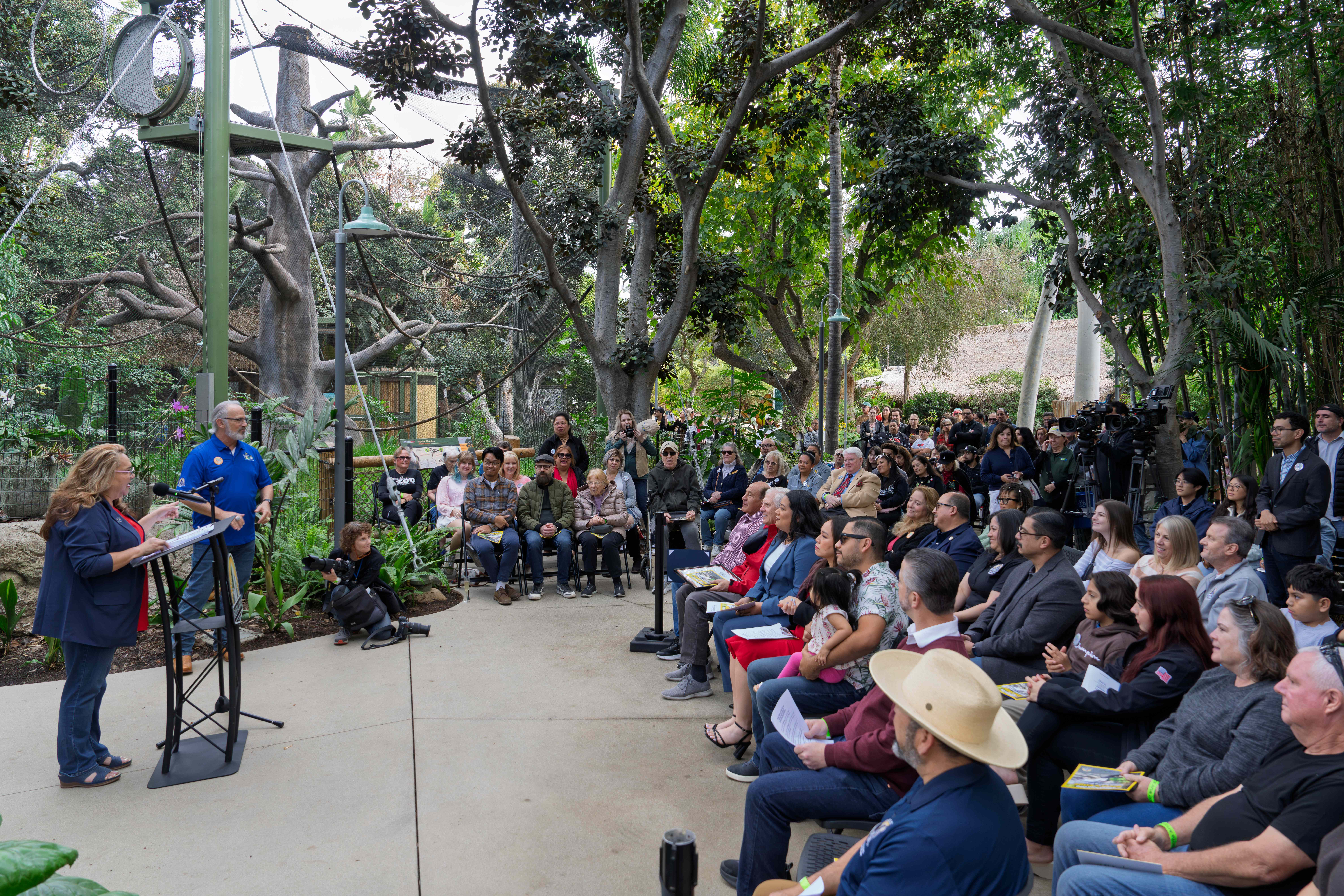 A woman speaks at a podium in front of a seated crowd outdoors with trees in background.